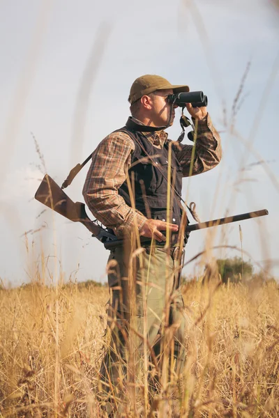 Man hunter with shotgun looking through binoculars in forest