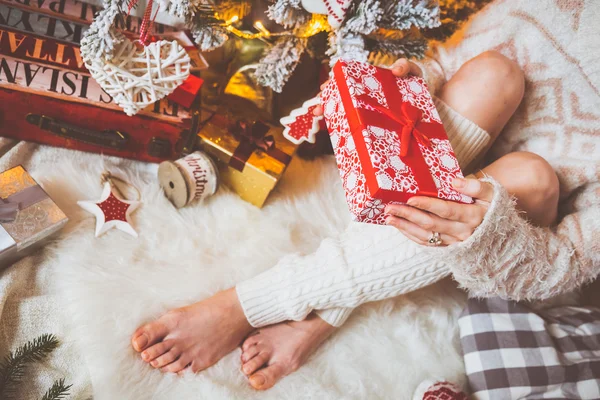 Young pretty woman in warm cozy clothes is sitting on the light wooden floor in her bright house near the christmas tree and holds gift box in her hands.