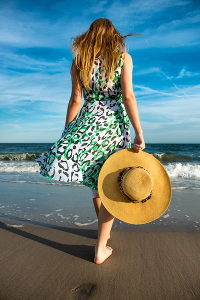 Young woman holding hat and walking on beach sand to the ocean. Photo of the woman from the back