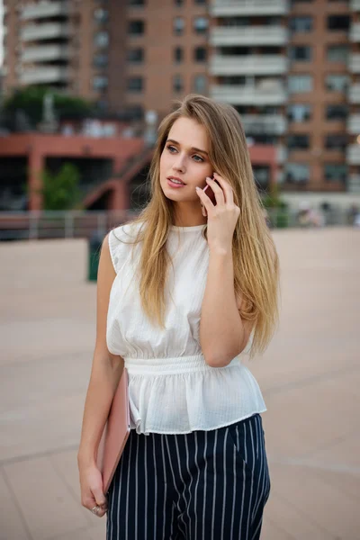 Beautiful young businesswoman using cell phone on city street. Beautiful blonde woman talking on the cell phone, holding laptop and walking on city street wearing business outfit.