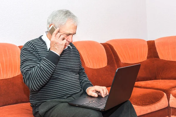 Senior businessman talking on cell phone and typing on laptop sitting on sofa. Concept photo of senior people and modern technology.