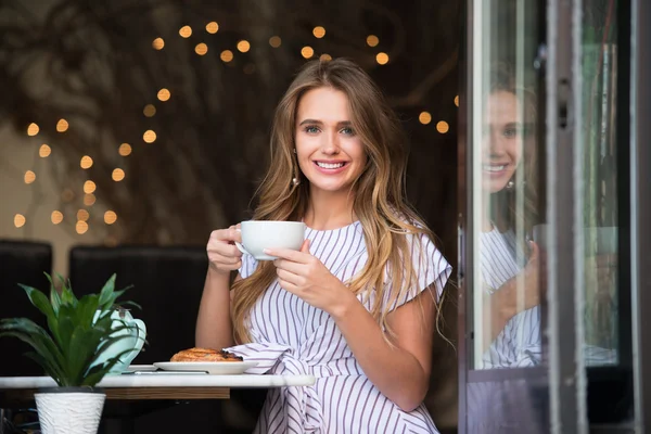 Beautiful smiling woman drinking coffee at the cafe and looking into the camera. Happy girl sitting in the cafe and having breakfast with coffee and croissant.