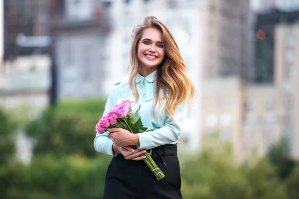 Beautiful business woman holding roses flowers bouquet and smiling in the city. Cheerful girl with cute smile enjoying flowers present.