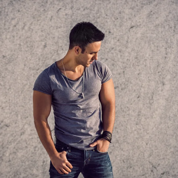 Portrait of fashion model man wearing grey t-shirt and jeans posing in front of the wall