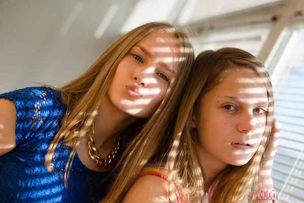 2 blond young women beautiful sisters or girl friends in blue dress having fun posing looking at camera against sun lighted rays through window blinds close up portrait image