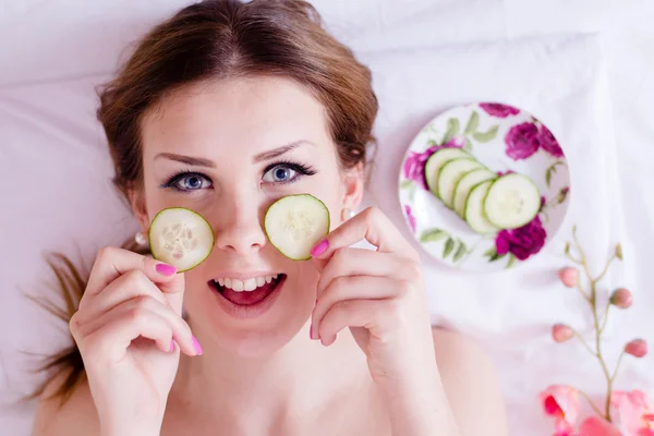 Green natural spa treatment: beautiful blond young woman having fun applying slices of cucumber to her face skin happy smiling & looking at camera on white background closeup portrait image