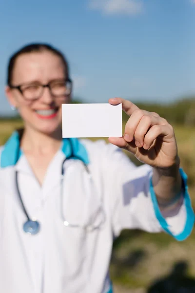 Closeup on business card: female doctor holding and showing blank card happy smiling & looking at camera on blue sky copy space background outdoors