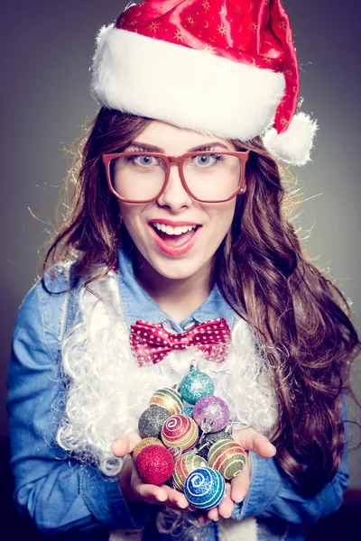 Funny hipster girl in super size eyeglasses wearing xmas santa hat and faux beard holding pile of Christmas tree decoration balls over olive copy space background