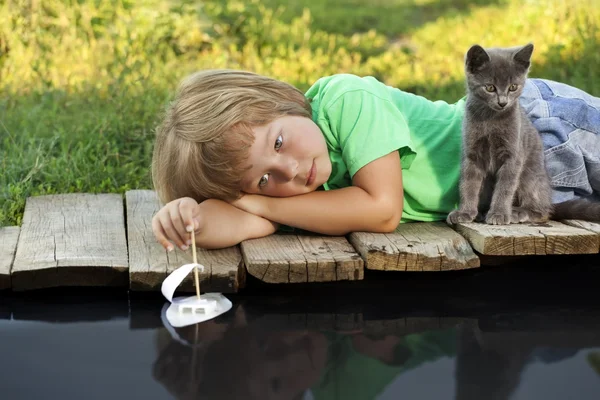 Boy and his beloved kitten playing with a boat from pier in pond