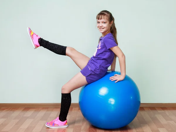 Little girl  is sitting on a blue fit ball, lifting the leg, smi