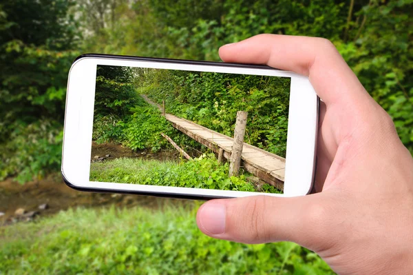 Hands taking photo landscape with a wooden bridge over a river with smartphone.