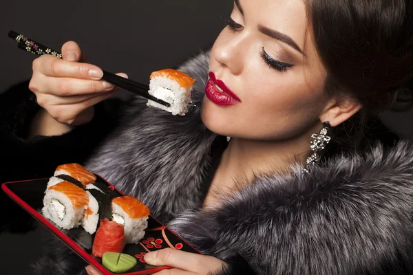 Beautiful young woman is eating sushi over black background