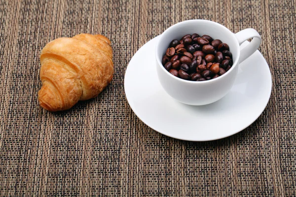 Coffee cup with a croissant and fresh coffee beans on a brown ba