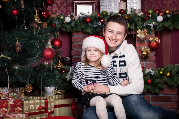 Father hugs daughter near the Christmas tree. The girls holds a small ball and there is a Santa Claus hat on her head. A fireplace behind them.