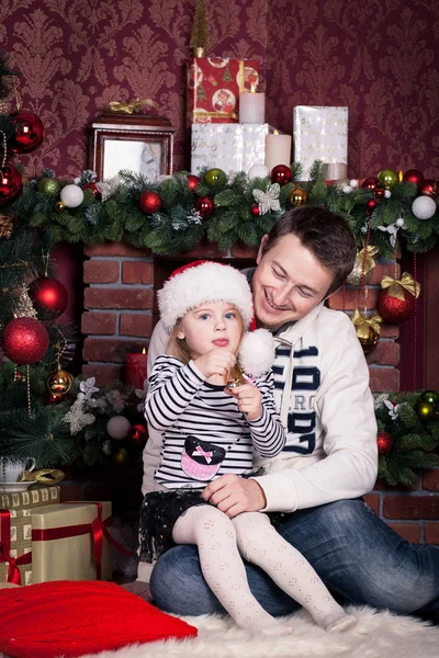 Father hugs daughter near the Christmas tree. The girls holds a small ball and there is a Santa Claus hat on her head. A fireplace behind them.