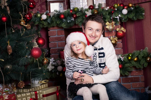 Father hugs daughter near the Christmas tree. The girls holds a small ball and there is a Santa Claus hat on her head. A fireplace behind them.