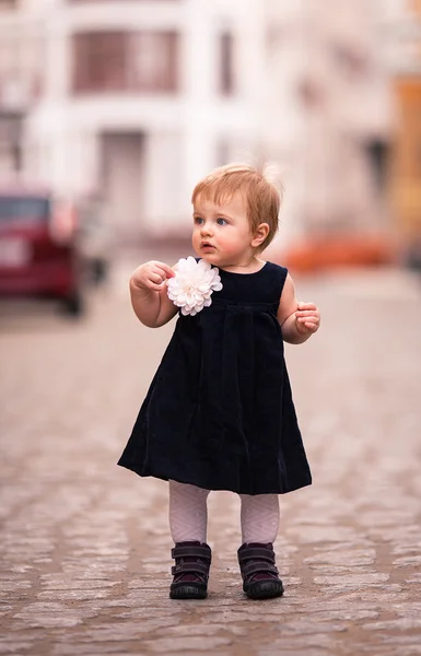A little baby girl in dark dress with big white flower stands on the street