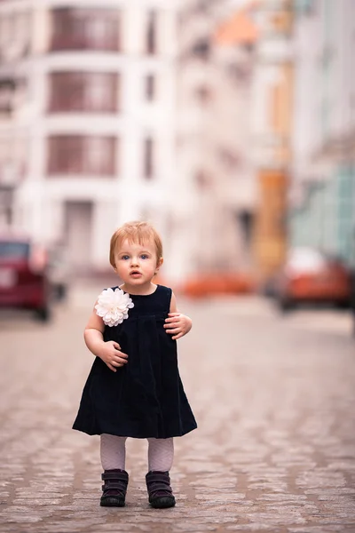 A little baby girl in dark dress with big white flower stands on the street