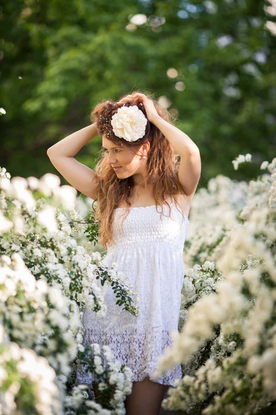 Charming young lady lifts up hel long curly dark hair in spring garden full of white flowers