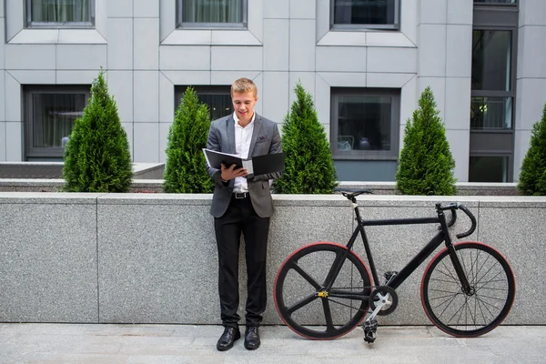 A young businessman standing on the steps of an office building, with a folder of papers and bike