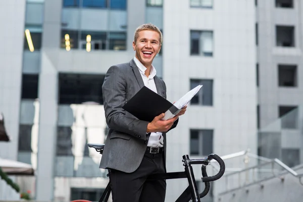 A young businessman standing on the steps of an office building, with a folder of papers and bike