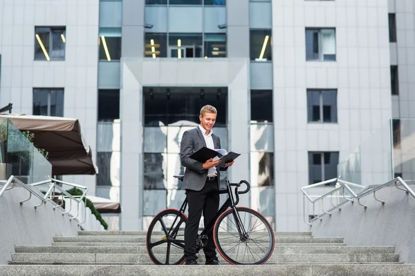 A young businessman standing on the steps of an office building, with a folder of papers and bike