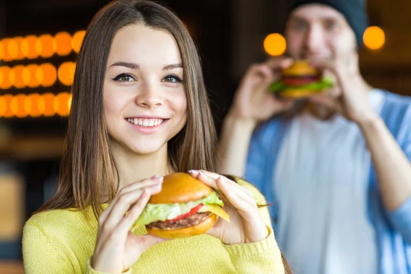 Man and woman eating burger. Young girl and young man are holding burgers on hands