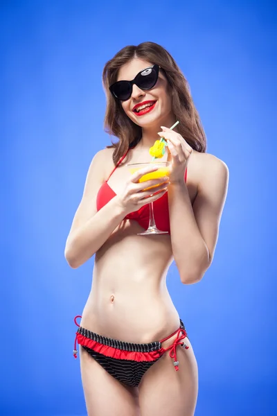 Slim bikini model in sunglasses holding drink against of blue background