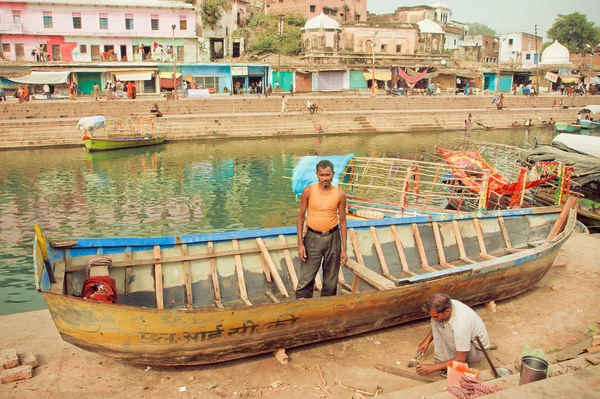 Repairmen repairing leaky fishing boat on river banks of indian city with historical ghats and houses