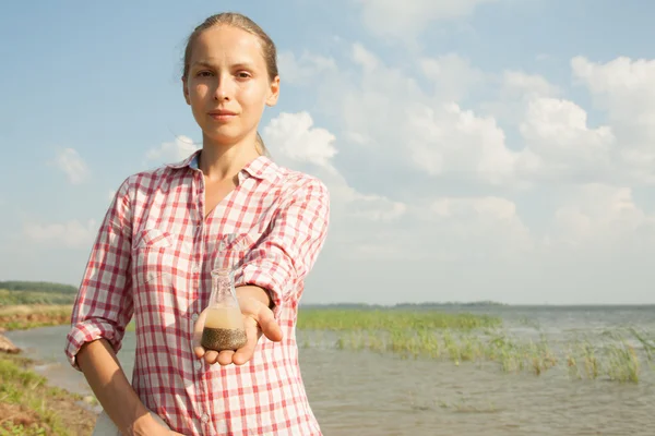 Water Purity Test. Woman holding a chemical flask with water, lake or river in the background.