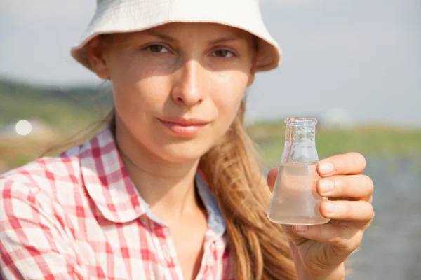 Water Purity Test. Woman holding a chemical flask with water, lake or river in the background.