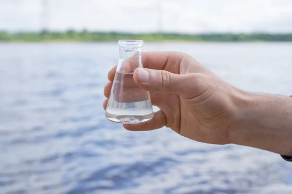 Hand holding a chemical flask with water, lake or river in the background.