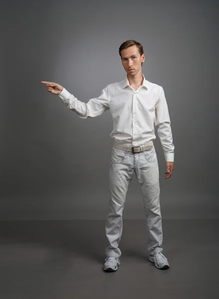 Young man in white shirt is pointing at something, standing on grey background