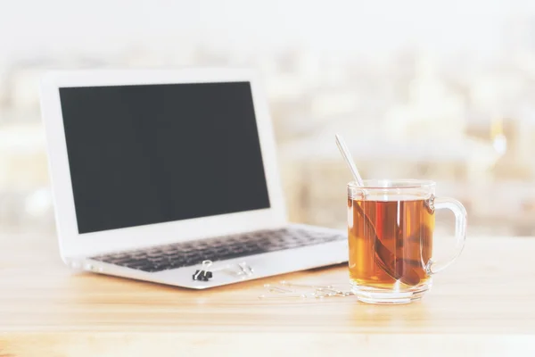 Sideview of wooden desktop with blank laptop screen and cup of tea on blurry city background. Mock up