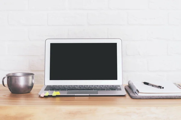 Front view of wooden desktop with blank black laptop screen, iron mug and other items on brick background. Mock up