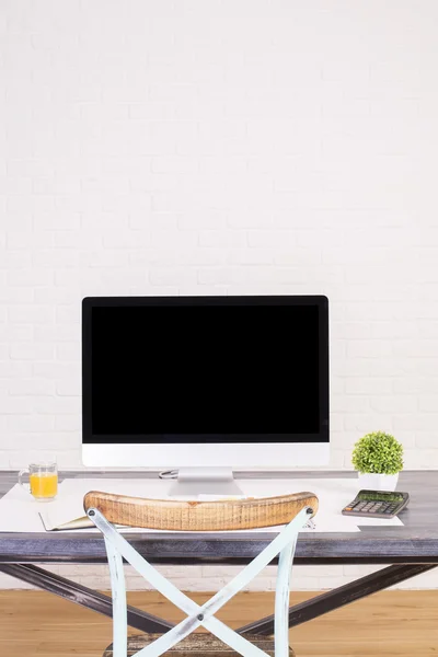 Designer tabletop with blank computer monitor and other items with an antique chair back showing. Wooden floor and white brick background.