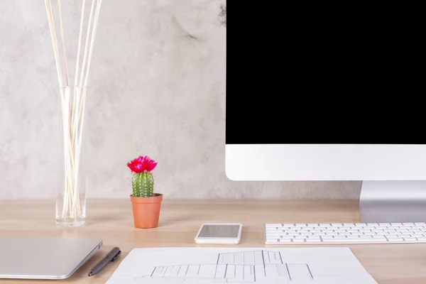 Creative desktop with blank computer monitor, cactus, smart phone, closed laptop, construction drawing and other items on concrete wall background. Mock up