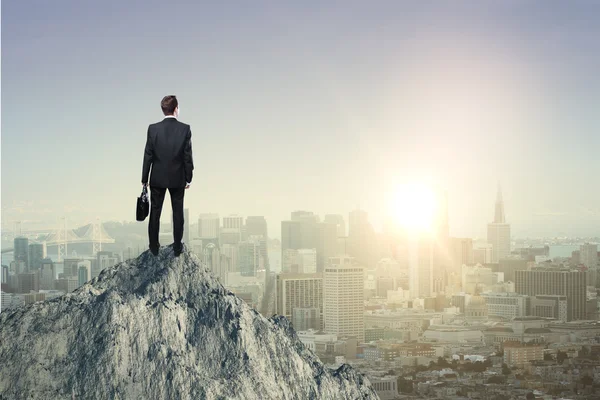 Research concept with businessman standing on mountain top and looking at sun setting over city
