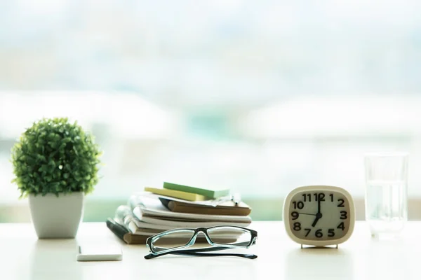 Closeup of bright desktop with glasses, pile of textbooks, decorative plant, water glass and clock on blurry background. Education concept