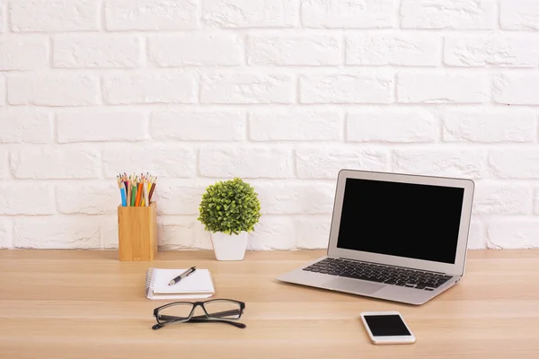Front view of creative designer desktop with blank laptop, cell phone, glasses, plant and stationery items on white brick wall background. Mock up