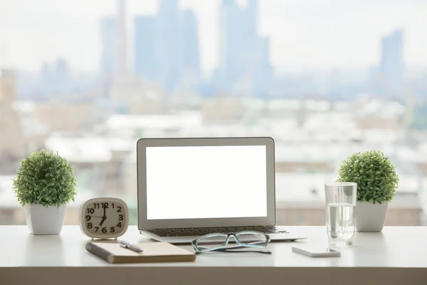 Creative windowsill workplace with blank white laptop computer, decorative plants, clock, water glass, smartphone, glasses and notepad with pen on blurry city background. Mock up