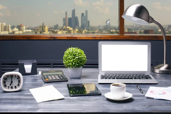 Creative hipster workplace with blank white laptop computer, tablet, coffee cup, table lamp, clock, calculator and other items on window with city view background. Mock up