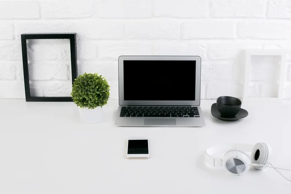 Front view of creative designer workplace with blank laptop screen, see-through picture frames, headphones, coffee cup, smartphone and plant on brick wall background. Mock up