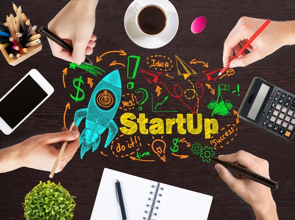 Startup concept with businesspeople hands drawing rocket ship sketch on wooden office desktop with blank smartphone, coffee cup, stationery and other items