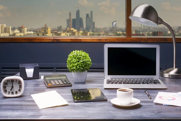 Creative hipster workplace with blank laptop computer, tablet, coffee cup, table lamp, clock, calculator and other items on window with city view background. Mock up