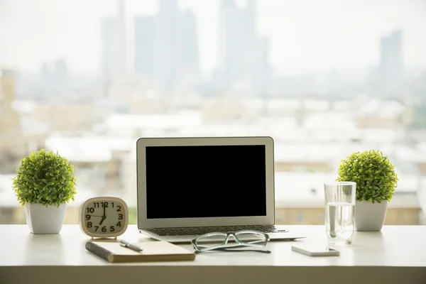 Creative windowsill workplace with blank laptop computer, decorative plants, clock, water glass, smartphone, glasses and notepad with pen on blurry city background. Mock up