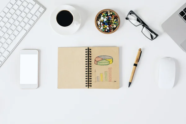 Top view of bright office desktop with blank white mobile phone, spiral notepad with drawn business chart, glasses, coffee cup, supplies, computer keyboards and mouse.