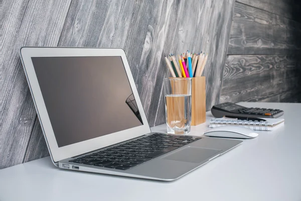 Side view of laptop on creative designer desktop with water glass, computer mouse, calculator and stationery items on wooden plank background. Mock up