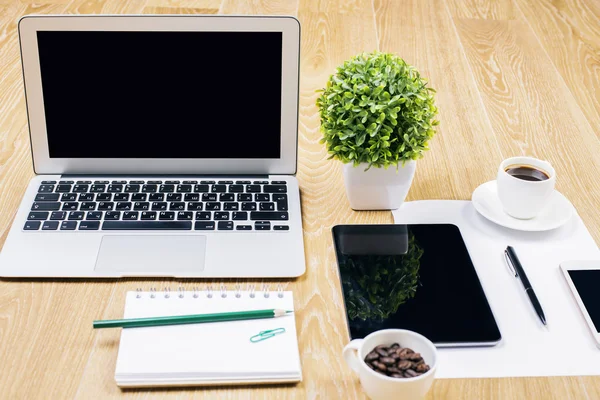 Front view and closeup of creative designer workplace with blank laptop, decorative plant, spiral notepad, coffee cups, tablet, smartphone and other items. Mock up