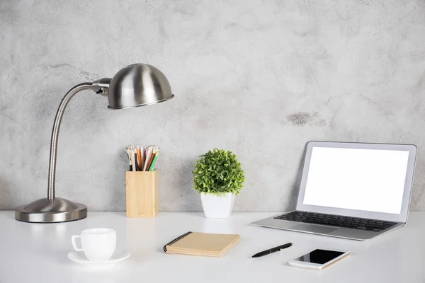 Creative designer desktop with blank white laptop computer, smartphone, spiral notepad, coffee cup, colorful pencils, plant and table lamp on concrete wall background. Mock up